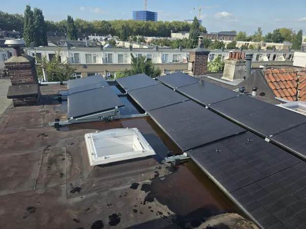 Zwarte zonnepanelen op schuine steunen op een plat bitumen dak tussen schoorstenen, met uitzicht over de stad